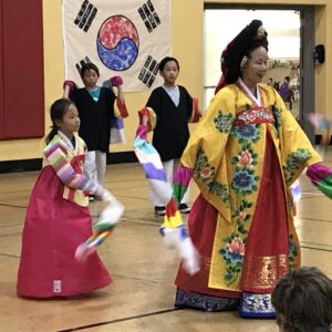 Korean woman in a hanbok with children in traditional Korean dress dancing behind her.